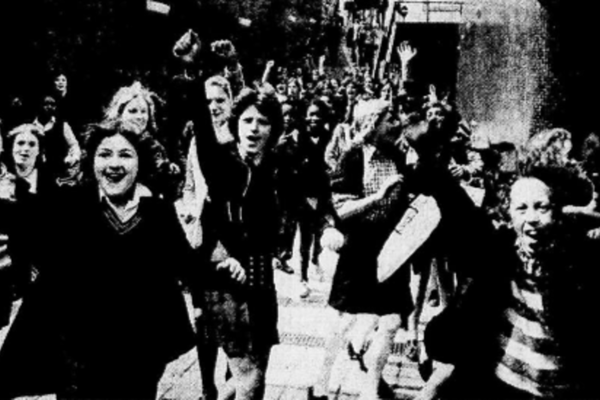 A group of young, school age girls running towards the camera in their school uniform. Their arms are raised in the air; some have their hands in a fist. Facial expressions range from happy and elated to determined and focused.